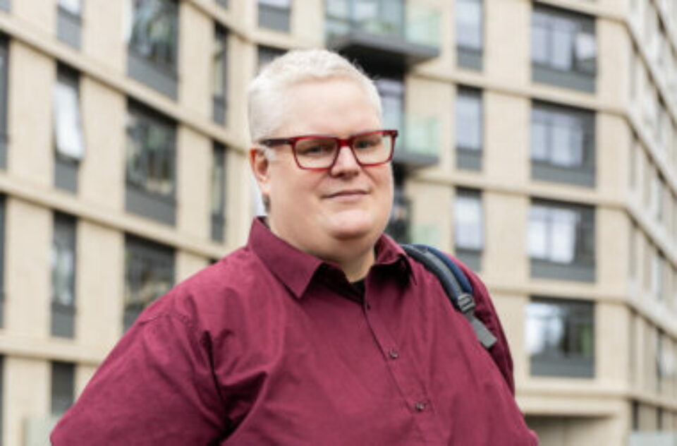 Hugo is a white man with short grey hair and glasses. He is wearing a deep red shirt and looking at the camera. He is standing in front of a canal.