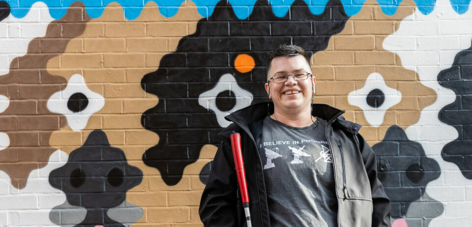 A man standing in front of a painted brick wall smiling at the camera. He is holding a cane and is wearing glasses, a black jacket and a grey t-shirt.