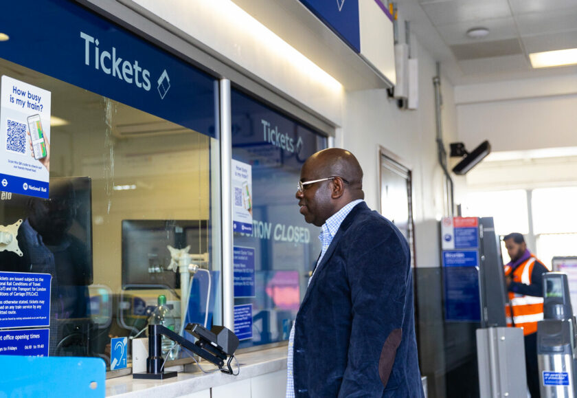 A black man wearing a blue velvet jacket stands in front of a Tickets Window in a London Underground station.