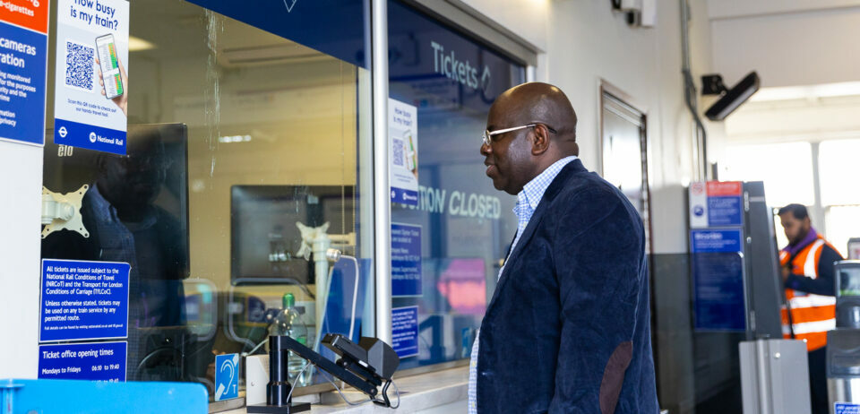 A Black man wearing a blue velvet jacket stands in front of a Tickets window in a London Underground station.