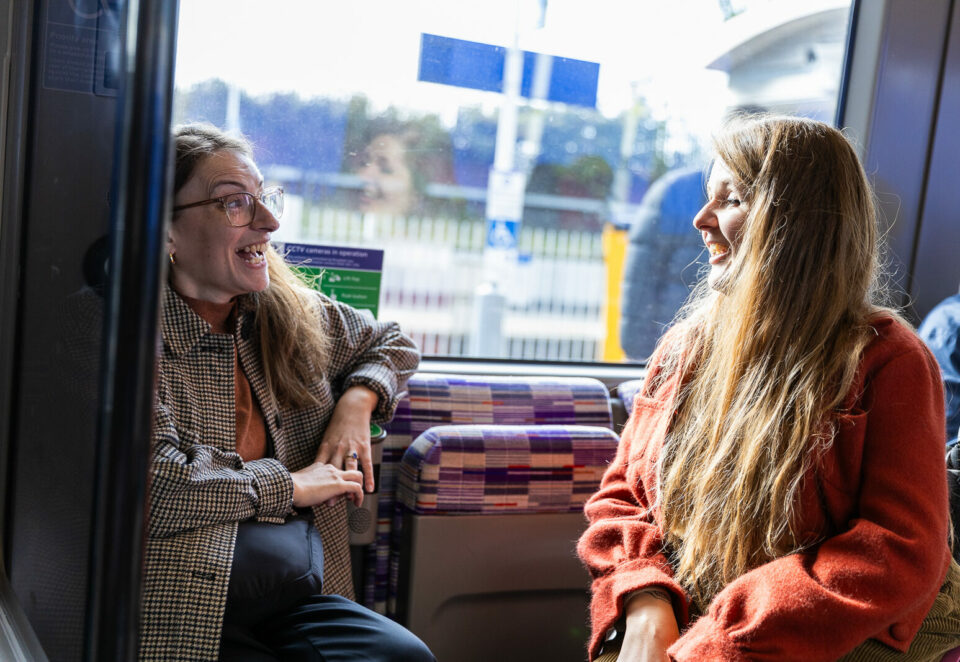 Two white women with long brown hair talking to at each other and smiling on a train. One of them is sitting on a fold-down seat and the other is using a wheelchair.