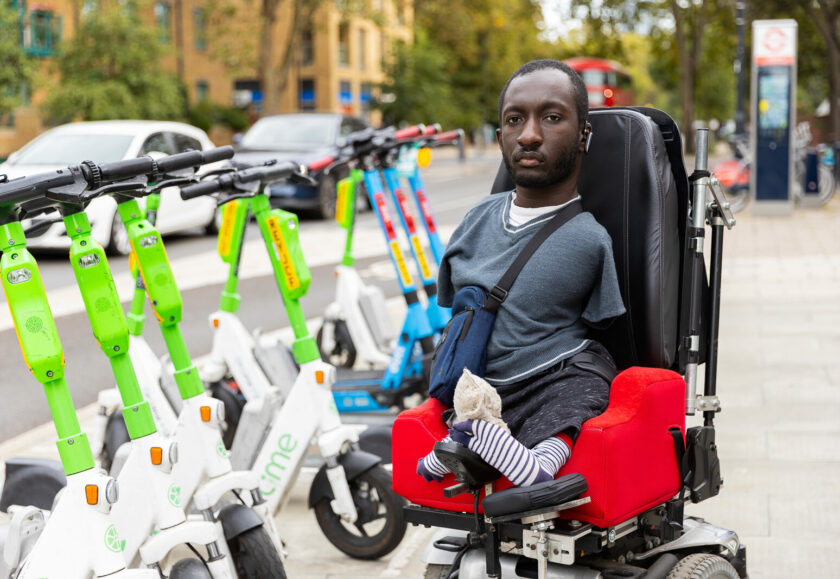 A Black man with upper and lower limb differences sits in an electric wheelchair. He has short brown hair and a beard, and is wearing a blue t-shirt and bag. On his left is a row of e-scooters which are parked on the pavement.