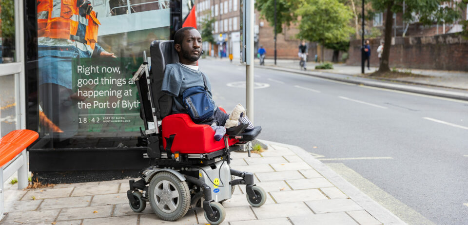A Black man with upper and lower limb differences sits at a bus stop in his electric wheelchair. He has short brown hair and a beard, and is wearing a blue t-shirt and bag.