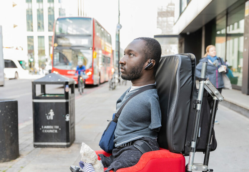 Side profile of a Black man with limb differences using a mobility chair on a city street. There is traffic visible on the road including a cyclist and a bus.