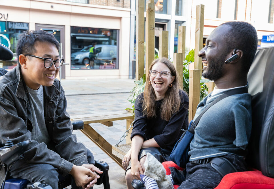 Three people are laughing together. On the right is a Black man with upper and lower limb differences who uses an electric wheelchair. He has short brown hair and a beard, and is wearing a blue t-shirt and bag. In the middle is a white woman sitting with her arms crossed. She has brown hair, wears glasses, and a black jumper. On the left is an Asian man who uses an electric wheelchair. He has dark hair, and wears glasses, a grey denim jacket, and jeans.