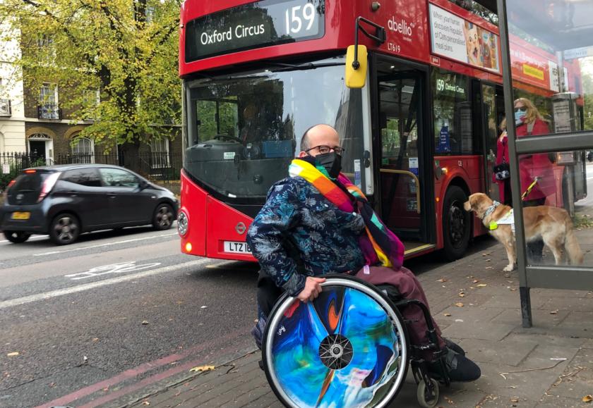 Two people are waiting at a bus stop. A number 159 bus is behind them. On the left is a white man in a blue and silver jacket with a rainbow scarf. He wears a black face mask and sits in a wheelchair with bright blue wheels. On the right is a woman with blonde hair, wearing a blue facemask and pink coat. With her is a guide dog.