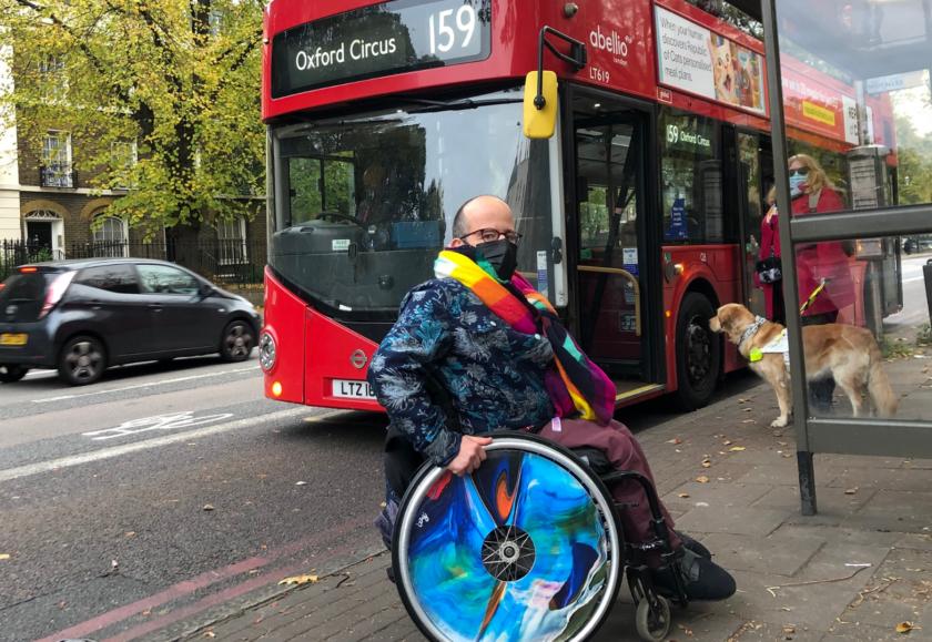 Two people are waiting at a bus stop. A number 159 bus is behind them. On the left is a white man in a blue and silver jacket with a rainbow scarf. He wears a black face mask and sits in a wheelchair with bright blue wheels. On the right is a woman with blonde hair, wearing a blue facemask and pink coat. With her is a guide dog.