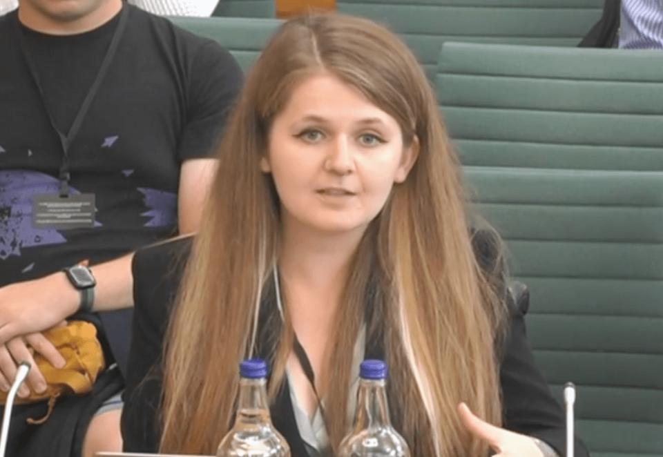 Katie, a white woman with long brown hair, is speaking in a Parliament committee room. She is wearing a shoulder-padded black power suit and looks serious.