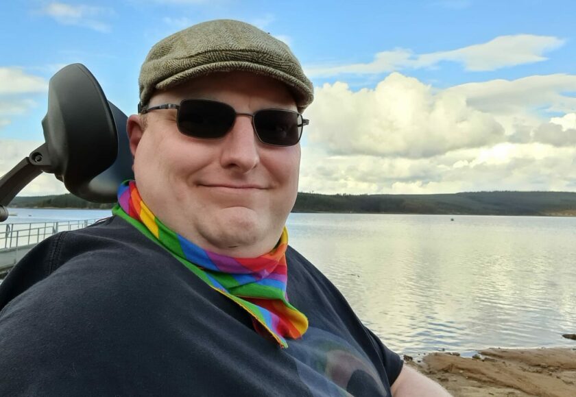 A smiling man wearing sunglasses, a hat and a rainbow coloured scarf looks into the camera. He is using a wheelchair and posing in front of a body of water.