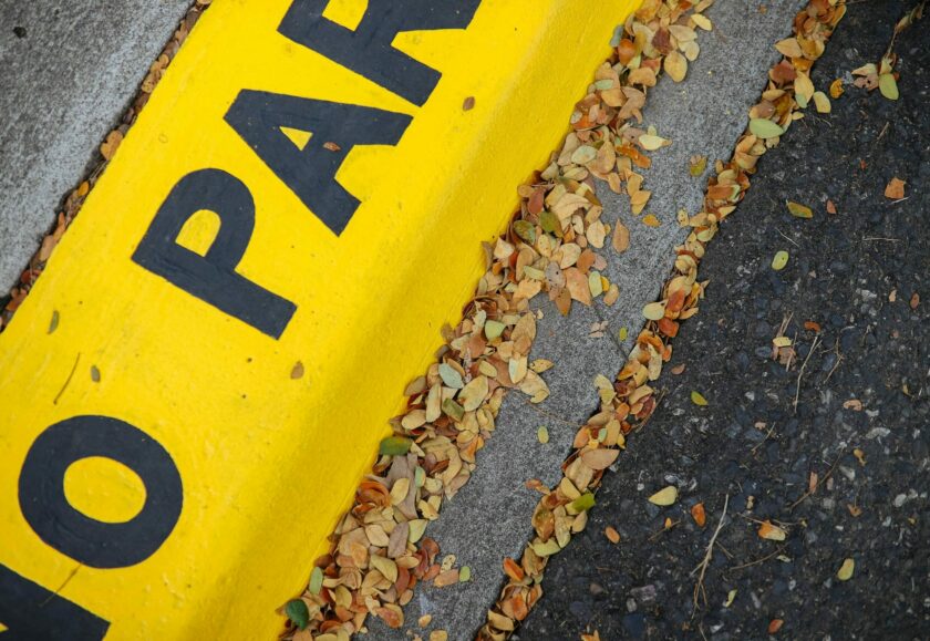 The kerb of a pavement with fallen leaves and yellow paint and black text which says 