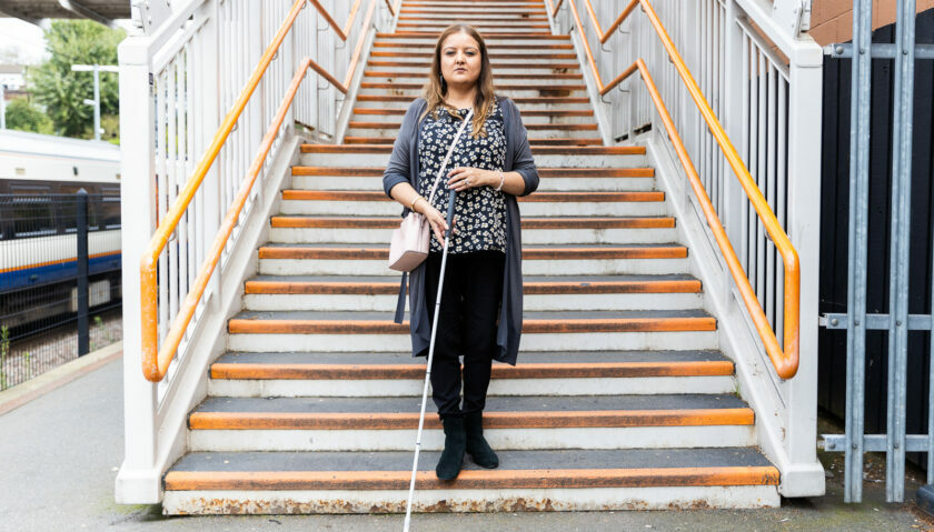 A woman holding a cane is standing on the bottom step of a staircase on a train platform. She looks defiant as she faces the camera. She wears a grey cardigan over a patterned blue top.