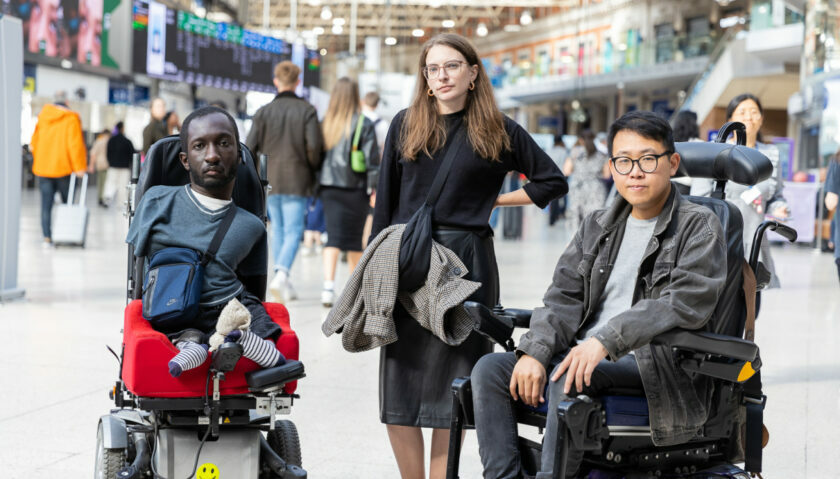 Three disabled people in a train station looking defiantly into the camera. On the left is a Black man with upper and lower limb differences who uses an electric wheelchair. He has short brown hair and a beard, and is wearing a blue t-shirt and bag. In the middle is a white woman standing with her arm on her hip. She has brown hair, wears glasses, and a black jumper. On the right is an Asian man who uses an electric wheelchair. He has dark hair, and wears glasses, a grey denim jacket, and jeans.
