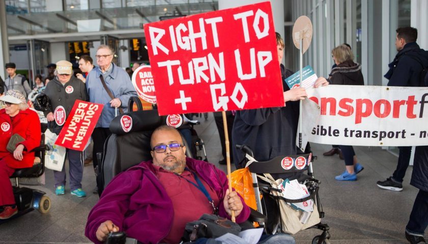 Ash Patel demonstrating at London Bridge against Southern Rail’s decision to suspend Turn Up and Go assistance (2017). Ash is an Asian man using a motorised wheelchair, and holds up a placard saying ‘Right to turn up + go’. He is outside the entrance of a train station, and there are several other protesters behind him, carrying signs that say ‘Southern betrayal’, ‘rail access now’, and ‘Transport for All, accessible transport is our right’.