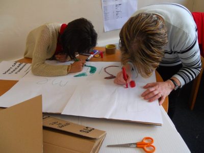 Two people are huddled over a table, each with different coloured markers in their hand, writing letters on a placard. There are other crafting tools like scissors and cardboard across the table. The faces of the two people are not visible. The person on the left has a dark brown bob and a tan cardigan, and the person on the right has short blond hair and a grey striped jumper.