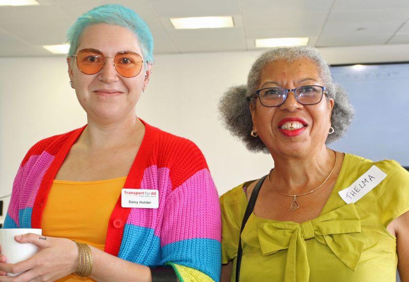 Two people stand next to each other, both smiling at the camera. On the left is a person wearing a bright cardigan, they have blue hair and tinted glasses. On the right is a woman with greying hair and a bright yellow top.