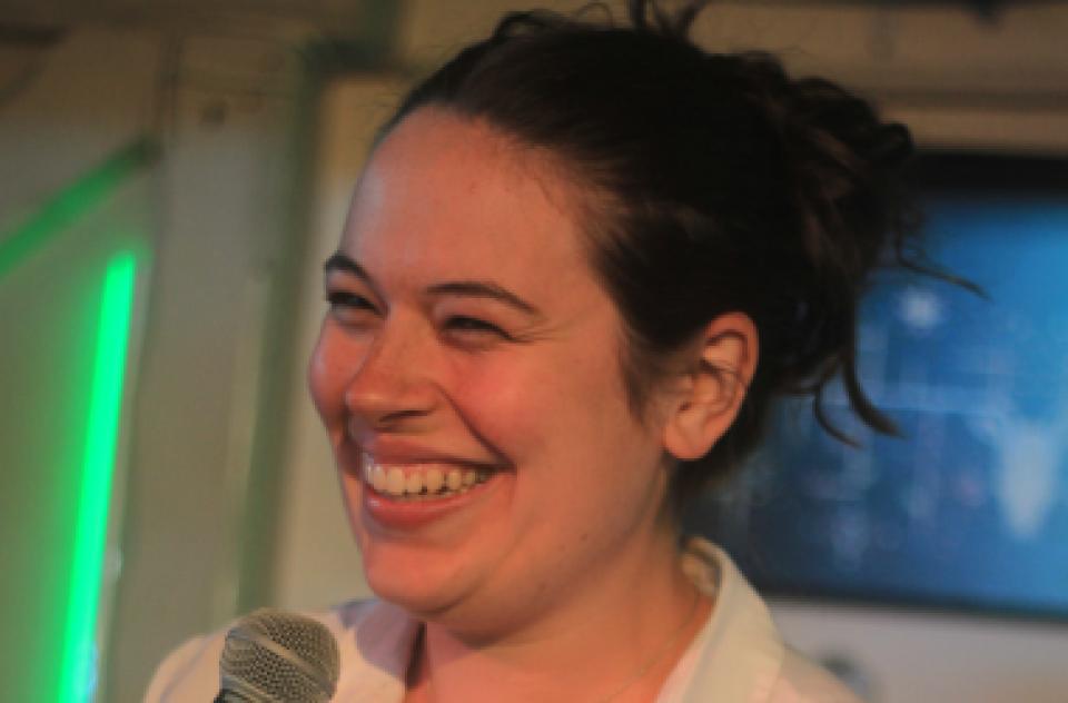 Camilla is a white woman with dark brown hair tied up in a bun and wearing a white shirt. She is standing with a microphone and smiling while looking away from the camera.